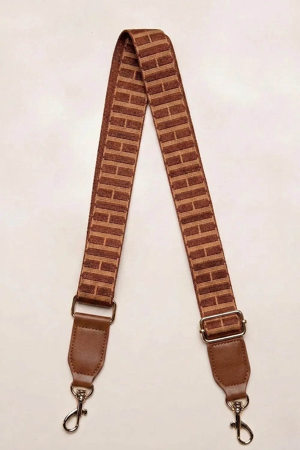 A brown fabric cognac tan strap with a H pattern, featuring brown leather end pieces and gold-toned metal swivel clips, laid flat on a light beige background.