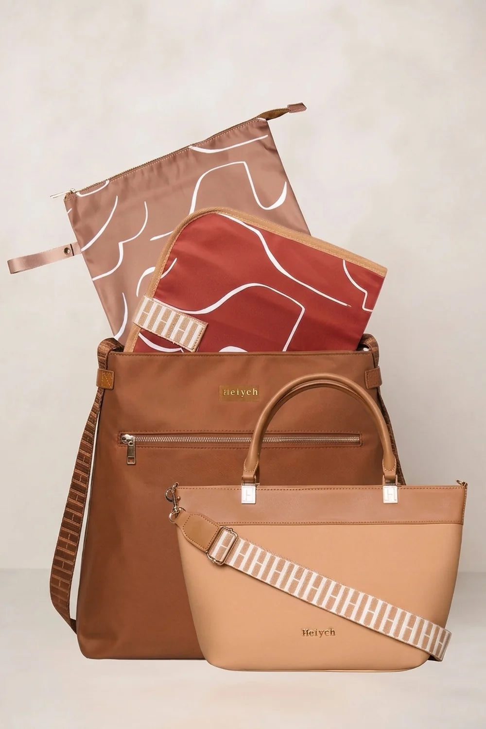 A coordinated travel bag set from Heyych, featuring a Tan Cognac tote bag, a rust-red zippered pouch with white abstract lines, and a dusty-rose reusable bag with a white line pattern, all arranged on a light beige background.