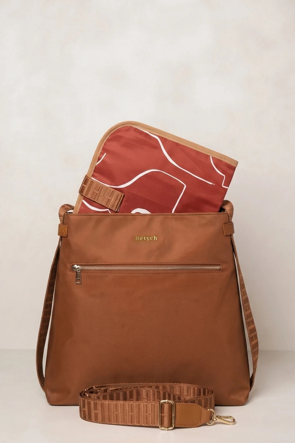 A cognac tan nylon shoulder bag with a matching brown leather strap, shown with a red and white patterned zippered pouch partially tucked inside. The brand name 'satch' is visible in gold lettering on the front.