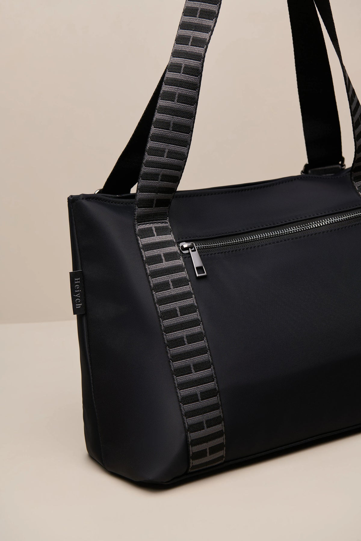 A black nylon duffel bag with a distinctive black-and-gray brick-patterned shoulder strap, shown on a neutral beige background.