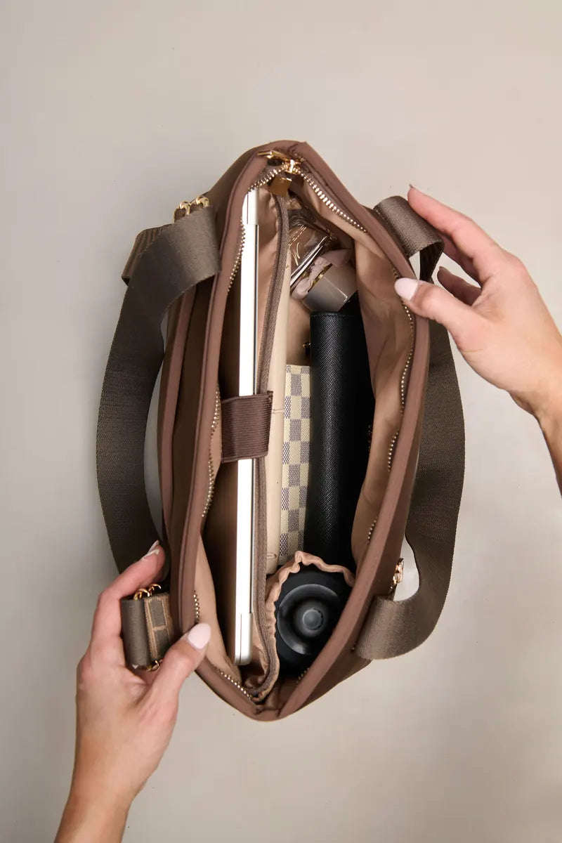 A brown chocolate shoulder bag is shown open with hands holding it, revealing a laptop, notebook, and other accessories inside. The bag has a wide shoulder strap and gold-tone zipper hardware, photographed against a plain light beige background.
