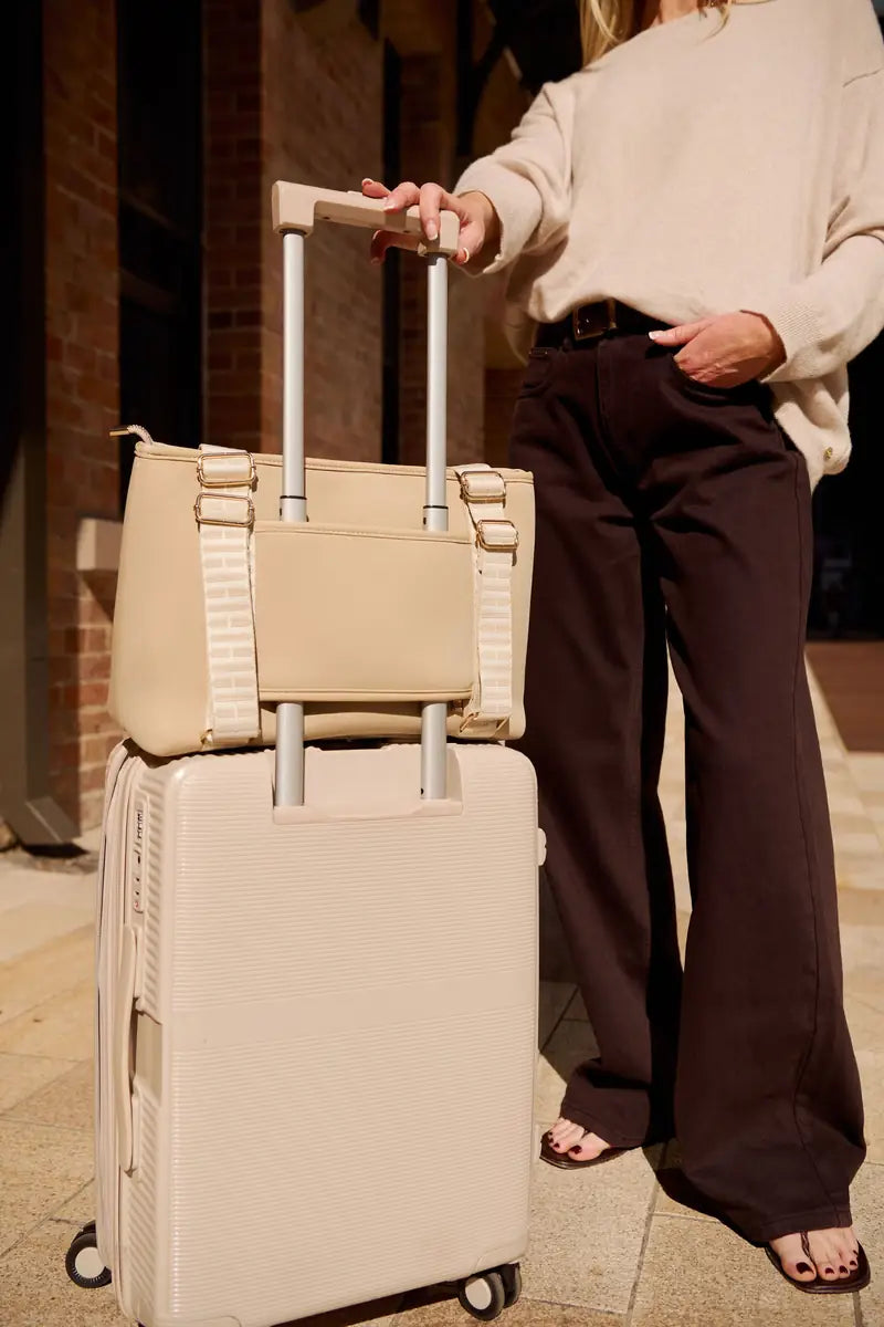 A woman in a cream sweater and dark brown wide-leg pants stands beside a beige hard-shell rolling suitcase with a matching taupe beige tote bag stacked on top, in an outdoor urban setting at night.