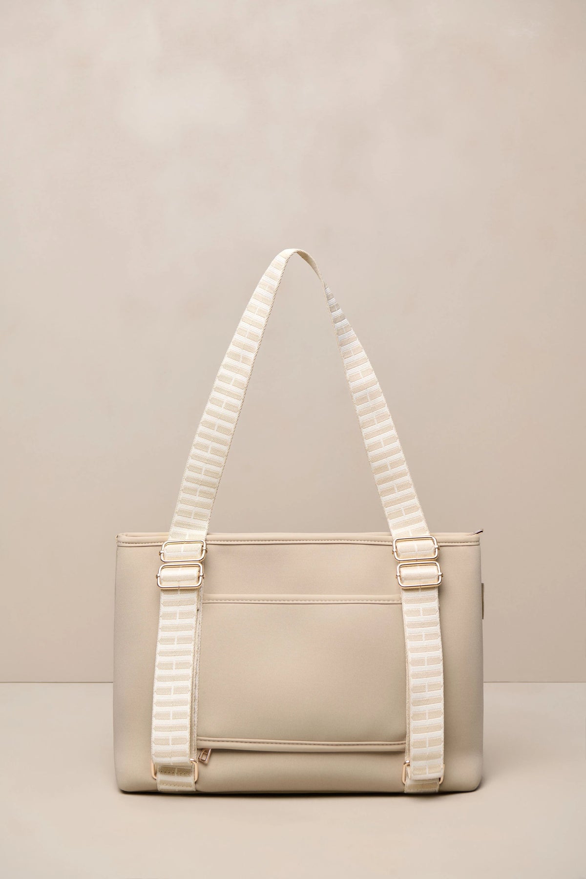 A structured taupe beige tote bag with wide, cream-and-beige striped shoulder straps and gold-tone hardware, set against a neutral beige background.