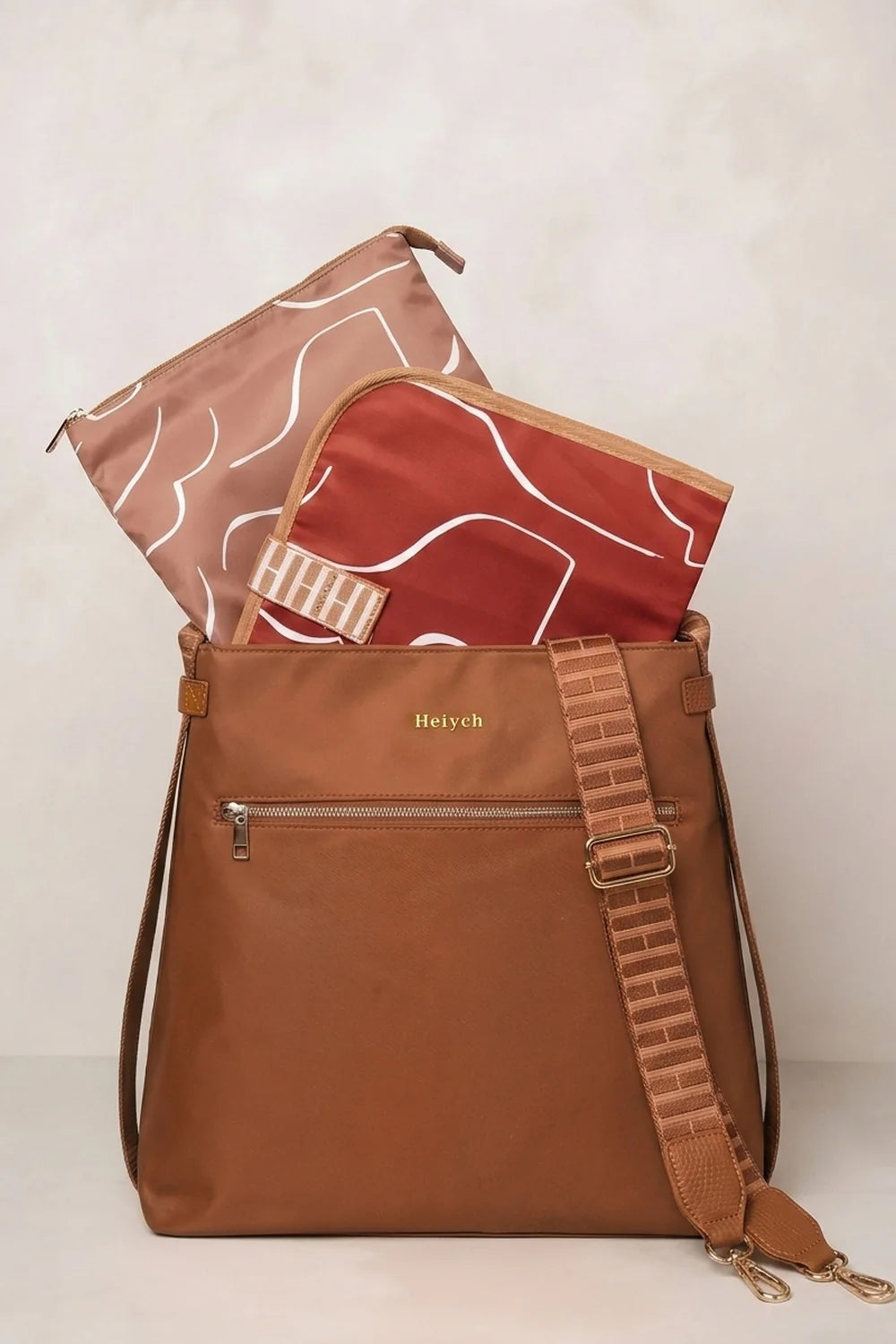 A cognac tan nylon messenger bag with a matching brown leather strap, shown with two patterned pouches partially tucked inside. The brand name 'Hestych' is printed in gold on the front pocket.