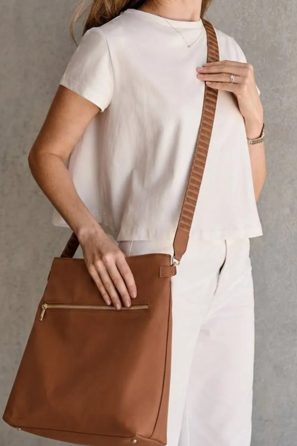 A woman wearing a white short-sleeve top and white pants carries a Tan Cognac crossbody bag with a matching brown strap against a textured gray wall.
