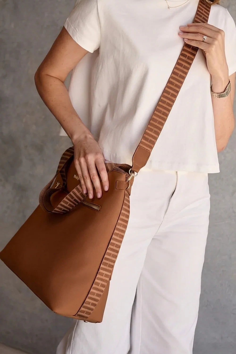 A woman wearing a white short-sleeve top and white pants carries a Tan Cognac crossbody bag with a wide brown-and-white striped strap against a gray wall.