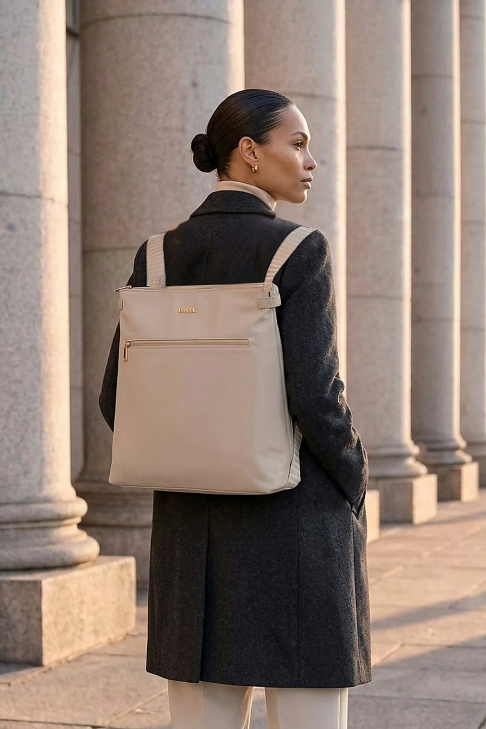 A woman in a dark gray coat stands in front of stone columns, carrying a large taupe beige backpack bag with a gold logo on the back.