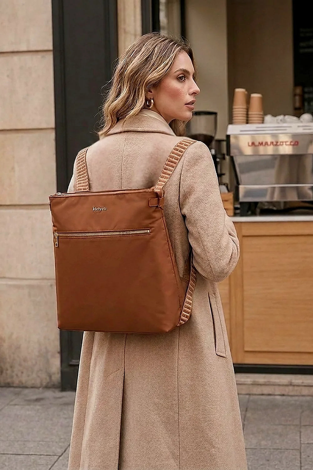 A woman in a beige coat is shown from behind wearing a cognac tan backpack with braided shoulder straps, set against a city street and cafe background.
