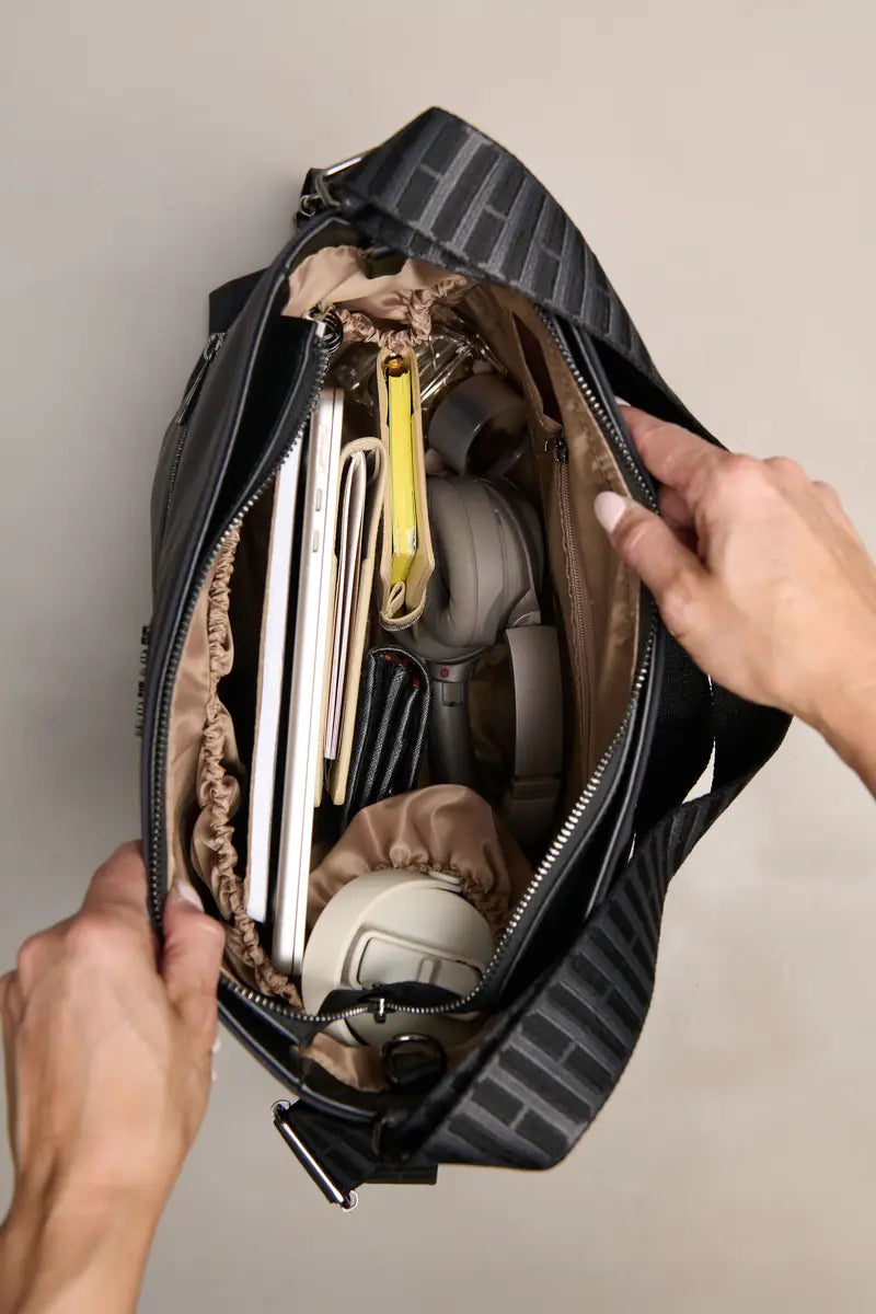 A top-down view of a black and gray striped duffel bag being opened by two hands, revealing its beige interior packed with travel accessories like a laptop, notebook, and headphones.