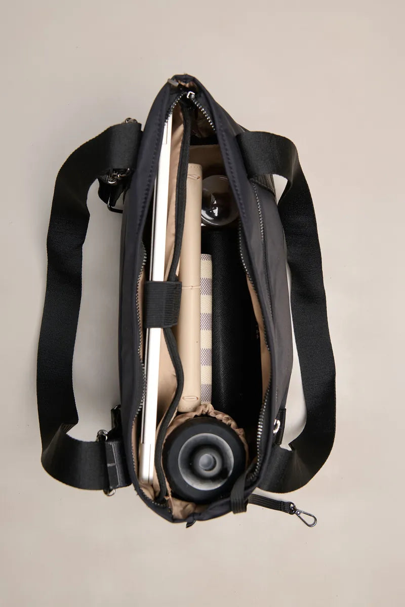 A black nylon shoulder bag shown from a top-down view, open to reveal its interior compartments containing a beige striped item and a black circular accessory, set against a solid light beige background.