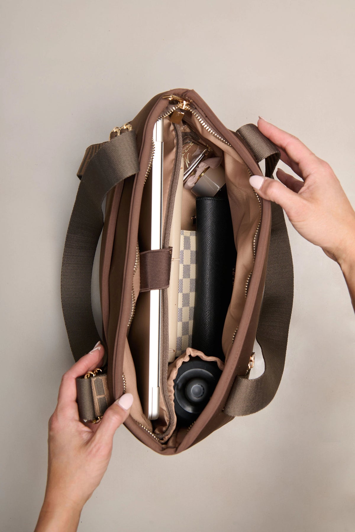 A brown chocolate shoulder bag is shown open with hands holding it, revealing a laptop, notebook, and other accessories inside. The bag has a wide shoulder strap and gold-tone zipper hardware, photographed against a plain light beige background.
