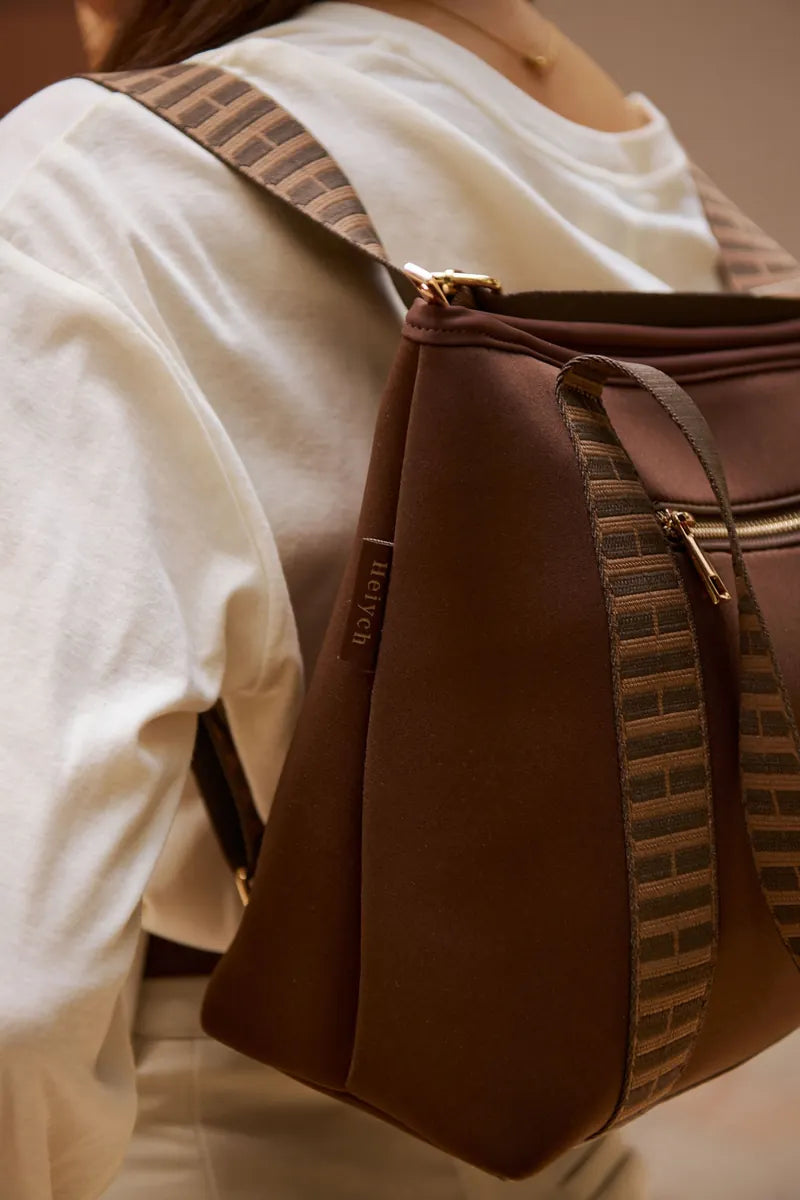 A woman wearing a white top carries a brown cognac shoulder bag with patterned brown and beige woven straps and gold-tone hardware.