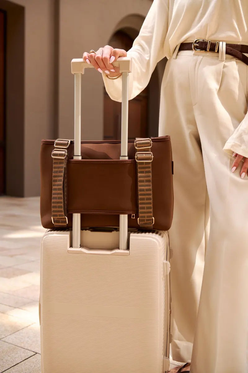 A woman in cream-colored clothing holds the handle of a cream hard-shell suitcase while carrying a brown chocolate luggage bag on top. The scene is set in a warm-toned indoor environment.