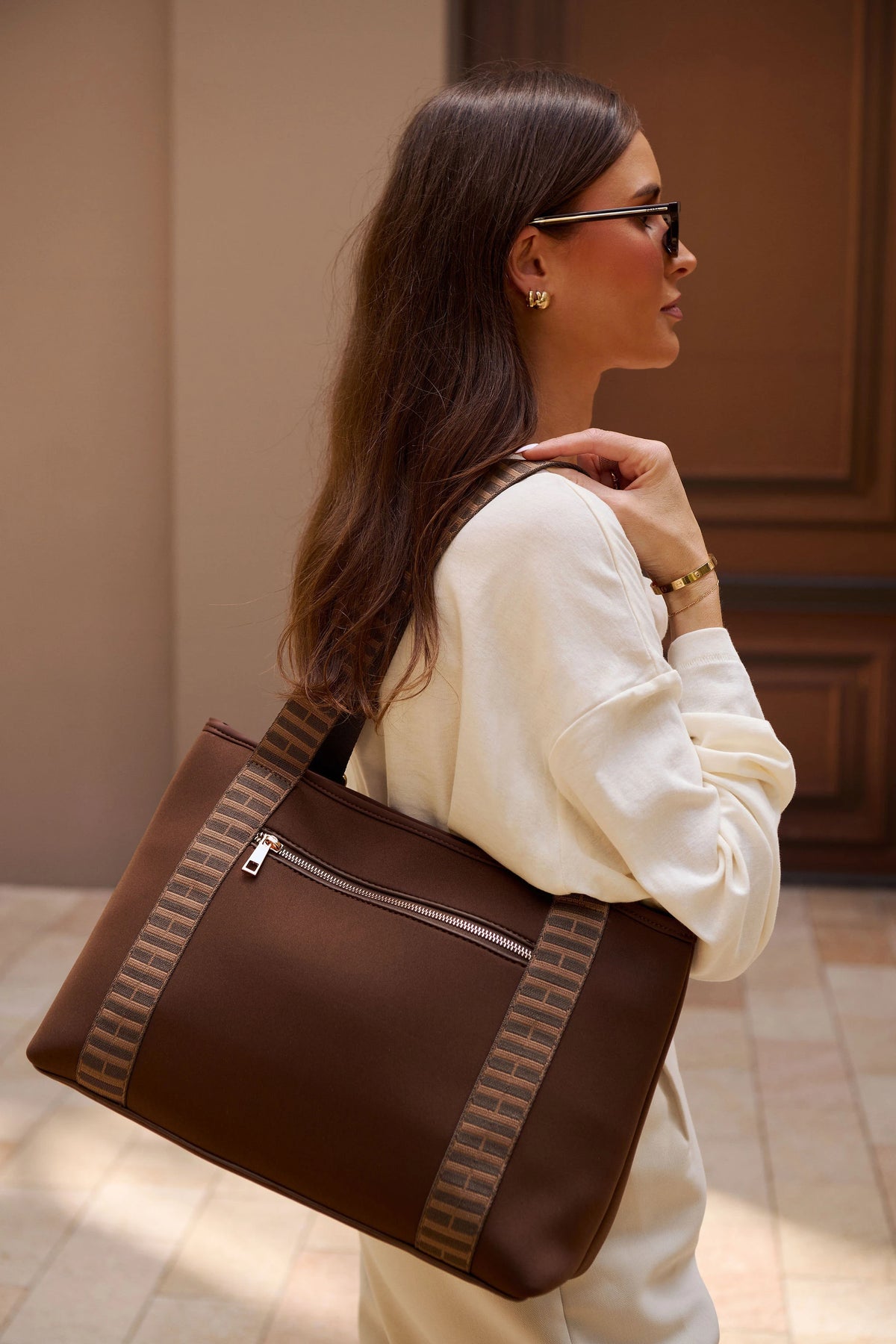 A woman in a cream sweatshirt and sunglasses carries a chocolate brown tote bag with patterned side straps in an indoor setting.