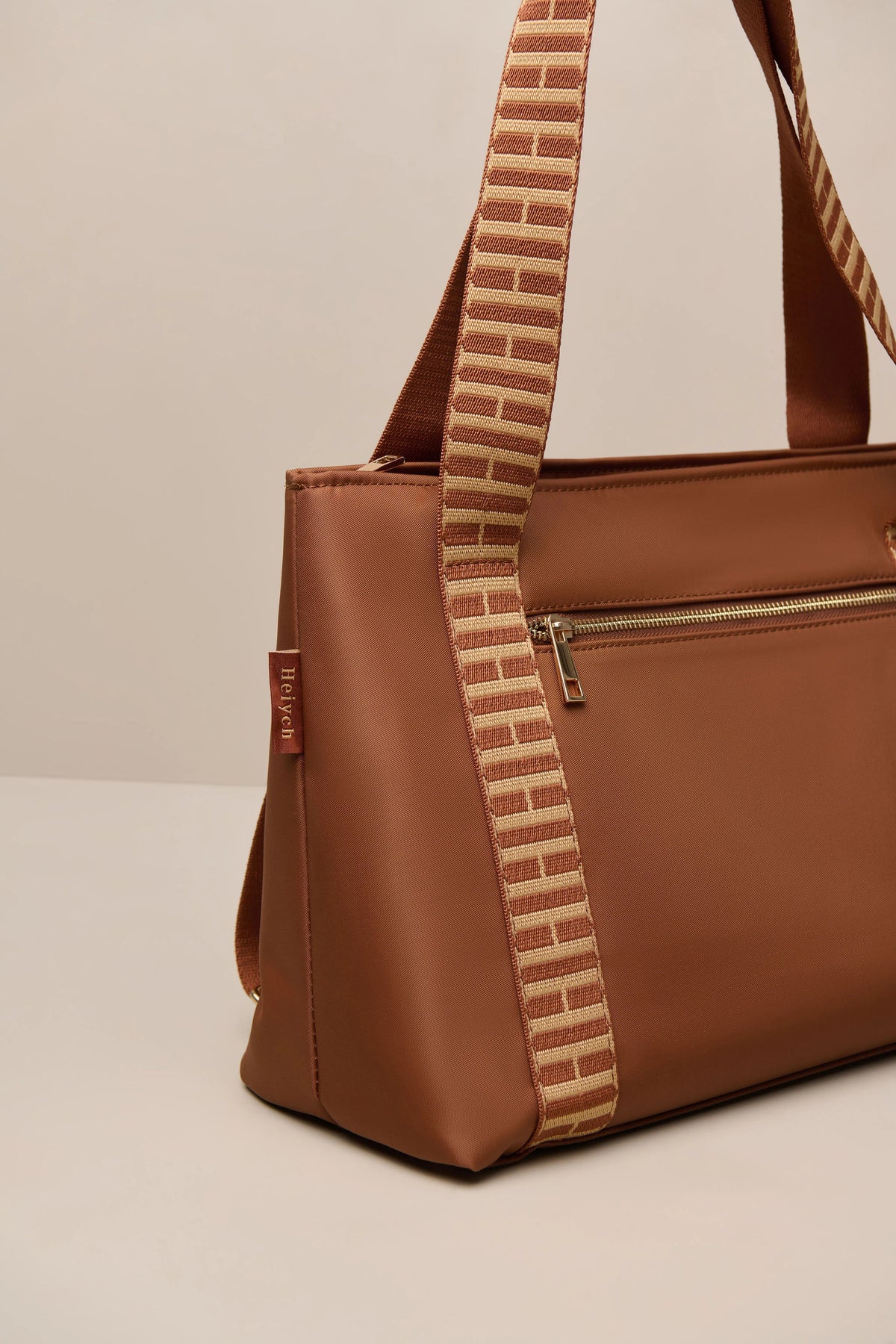 A brown cognac tote bag with a distinctive woven shoulder strap featuring a brick pattern in beige and brown, set against a neutral light beige background.