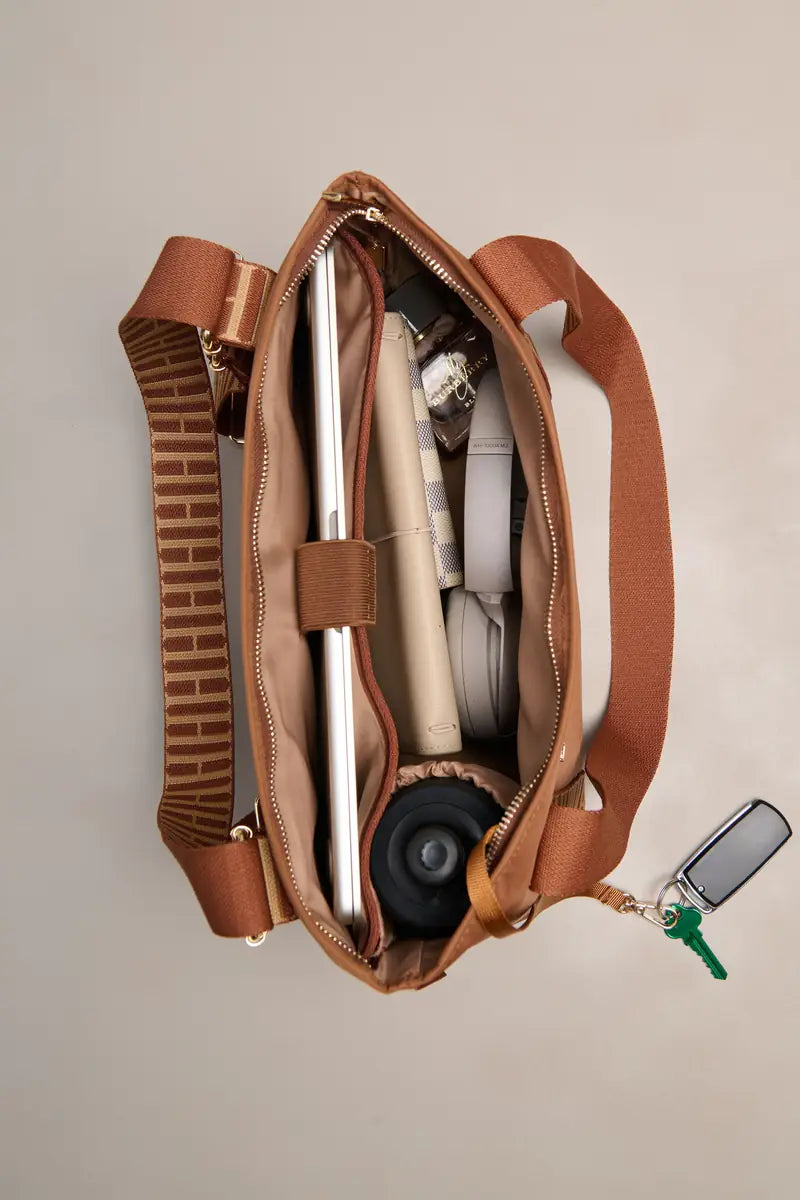 An open brown cognac nylon shoulder bag shown from above, packed with a tablet, notebook, headphones, and other accessories, on a light beige background.
