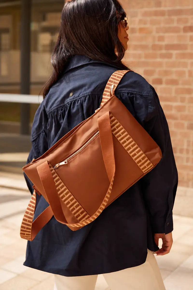 A woman wearing a navy blue jacket and beige pants is shown from behind, carrying a brown crossbody bag with patterned straps over her shoulder. The bag has a cognac brown body with beige and brown striped patterned straps and a silver zipper.