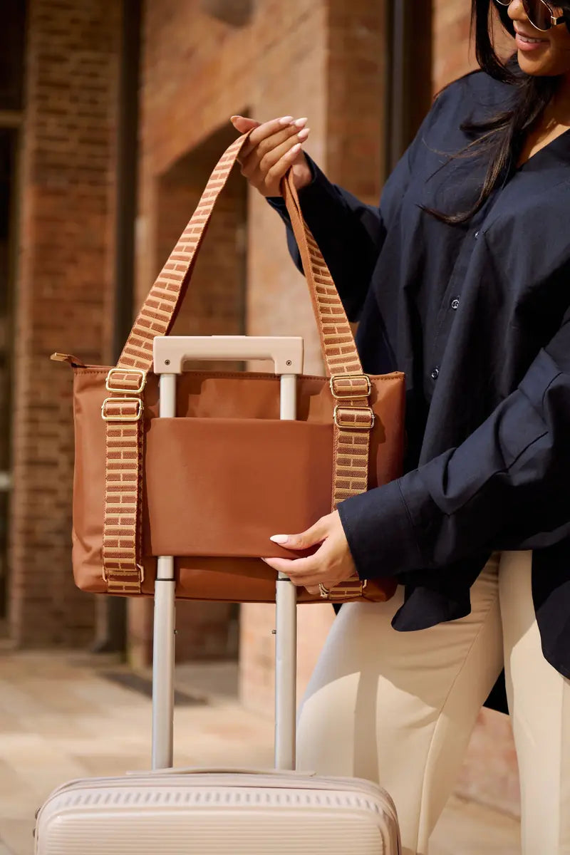 A woman in a navy shirt and cream pants holds a brown cognac and woven strap tote bag next to a light beige rolling suitcase in front of a brick building.
