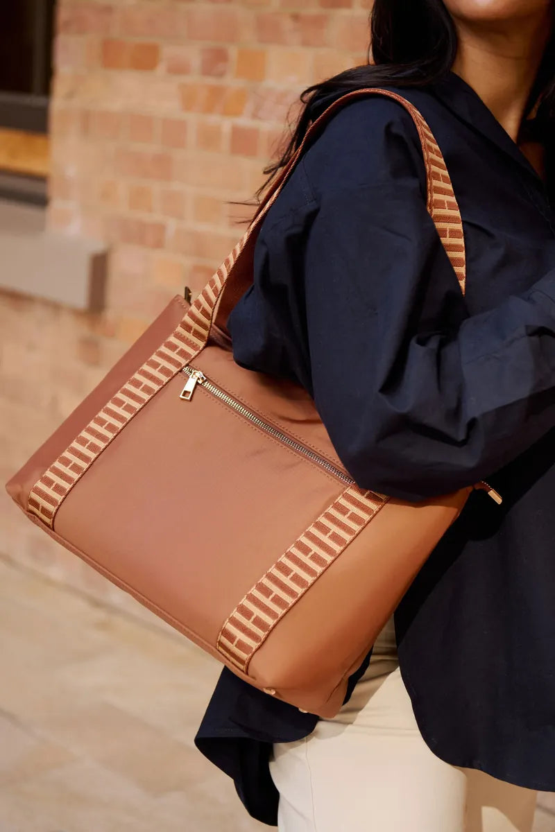 A person wearing a navy blue shirt and white pants carries a tan cognac shoulder bag with beige and white striped woven straps, set against a blurred brick wall background.