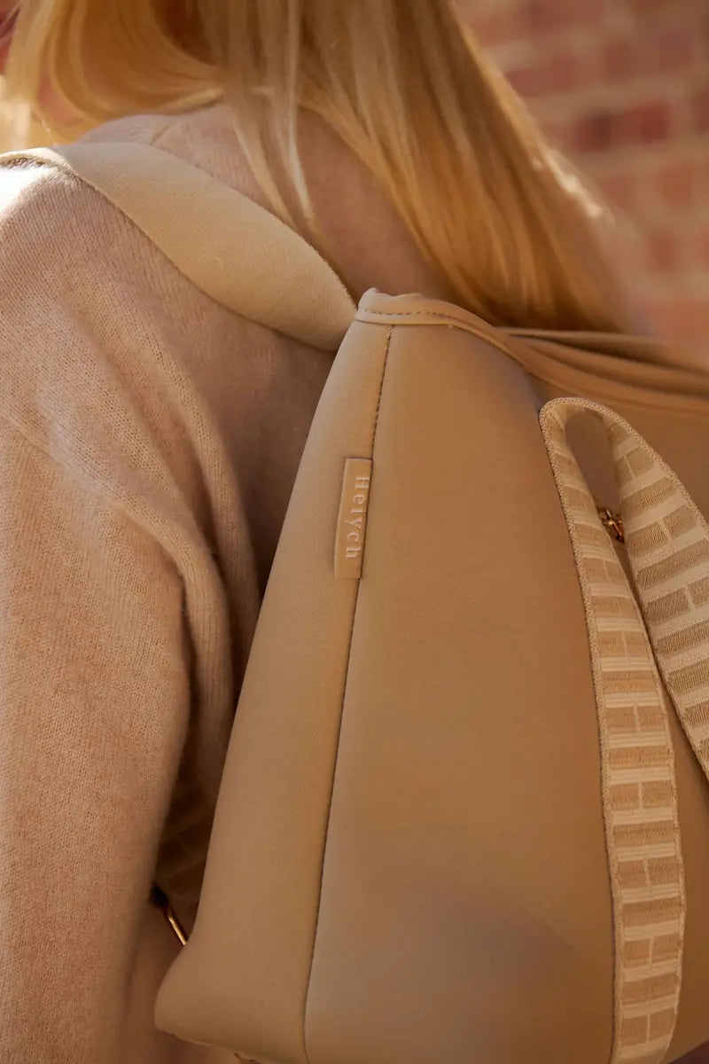 A close-up lifestyle shot of a model wearing a beige taupe shoulder bag with a matching beige knit strap, set against a soft-focus background of a brick wall and warm sunlight.