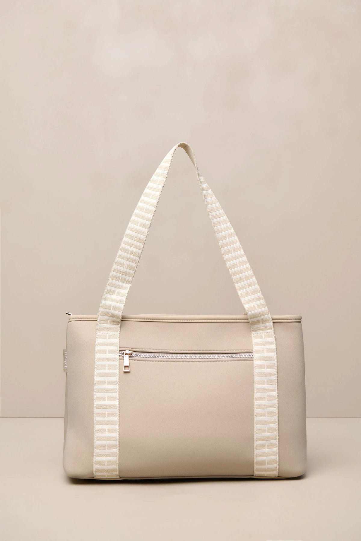 A structured beige taupe tote bag with cream and beige striped shoulder straps, featuring a front zippered pocket and a top zipper closure, set against a neutral beige background.