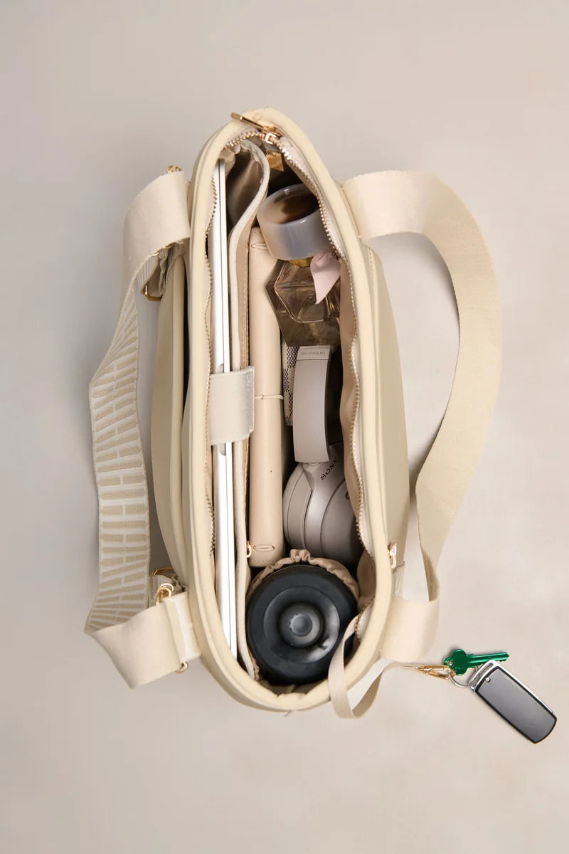 An open taupe-colored shoulder bag with a wide strap, shown from a top-down view on a light beige background. The bag is packed with various items, including a silver laptop, a pink pen, a white cord, and a drink bottle, with a small key fob hanging from the side zipper.